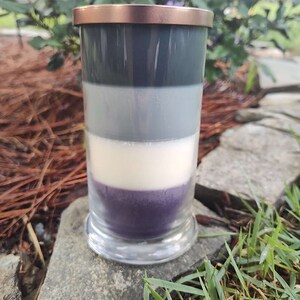 May include: A layered candle in a clear glass jar with a copper lid. The candle is made of four layers of wax: dark gray, light gray, white, and purple.
