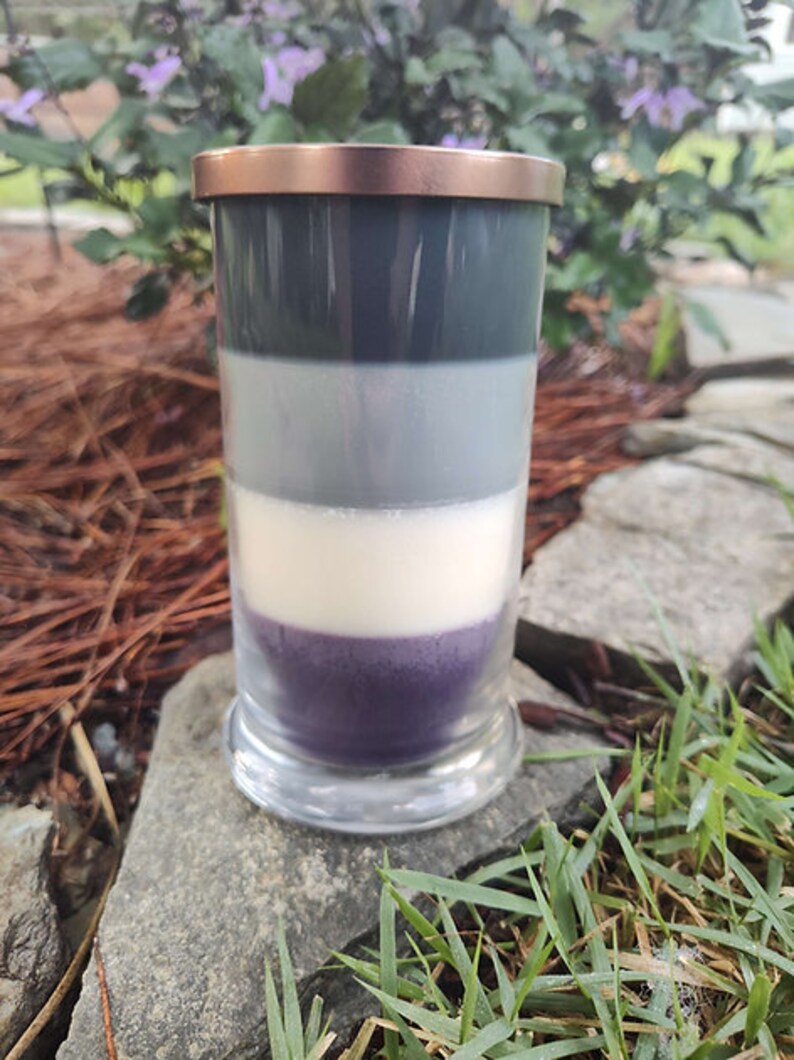 May include: A layered candle in a clear glass jar with a copper lid. The candle is made of four layers of wax: dark gray, light gray, white, and purple.