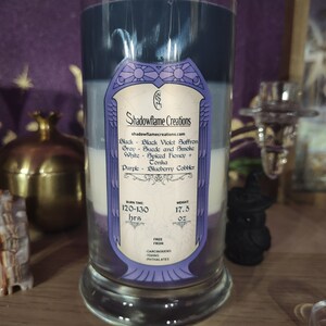 May include: A glass candle with four layers of wax in shades of purple, white, gray, and black. The label reads "Shadowflame Creations" and lists the scents: Black Violet Saffron, Suede and Smoke, Spiced Honey, and Blueberry Cobbler. The candle has a burn time of 120-130 hours and weighs 17.5 ounces. It is free from carcinogens, toxins, and phthalates.