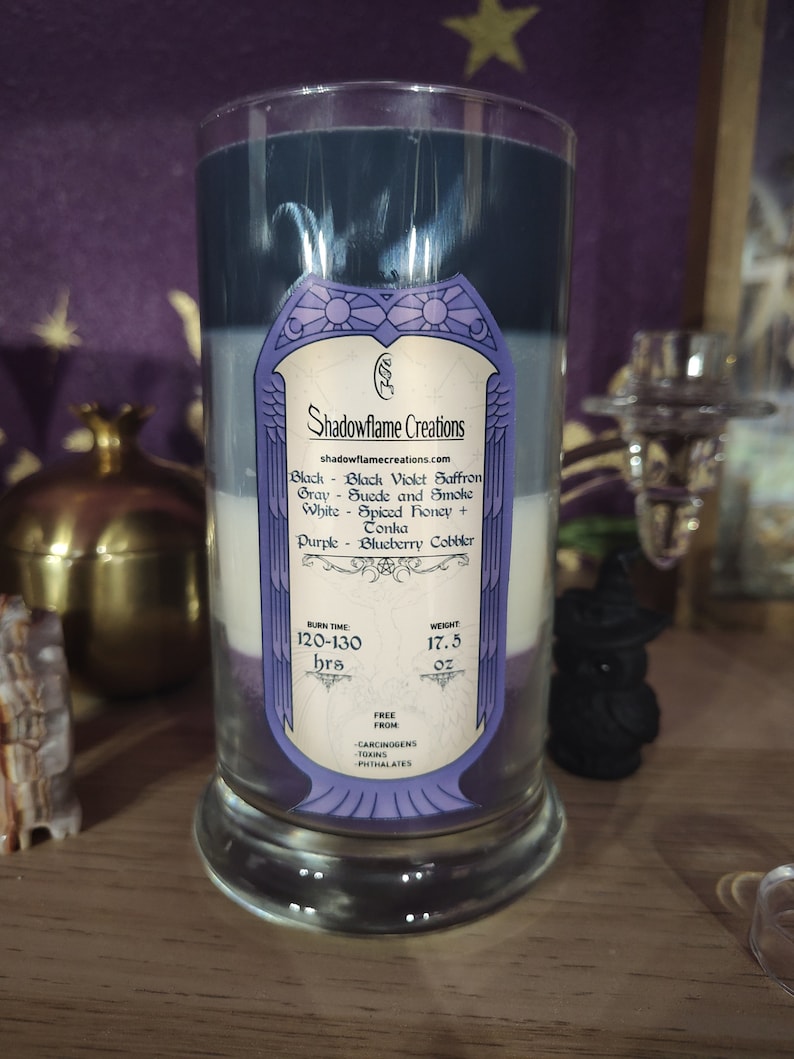 May include: A glass candle with four layers of wax in shades of purple, white, gray, and black. The label reads "Shadowflame Creations" and lists the scents: Black Violet Saffron, Suede and Smoke, Spiced Honey, and Blueberry Cobbler. The candle has a burn time of 120-130 hours and weighs 17.5 ounces. It is free from carcinogens, toxins, and phthalates.