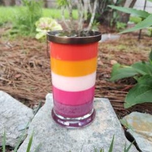 Op de afbeelding: Een gelaagde kaars in een glazen pot met een metalen deksel. De kaars is in de kleuren van de lesbische pride-vlag: roze, wit, oranje en rood.