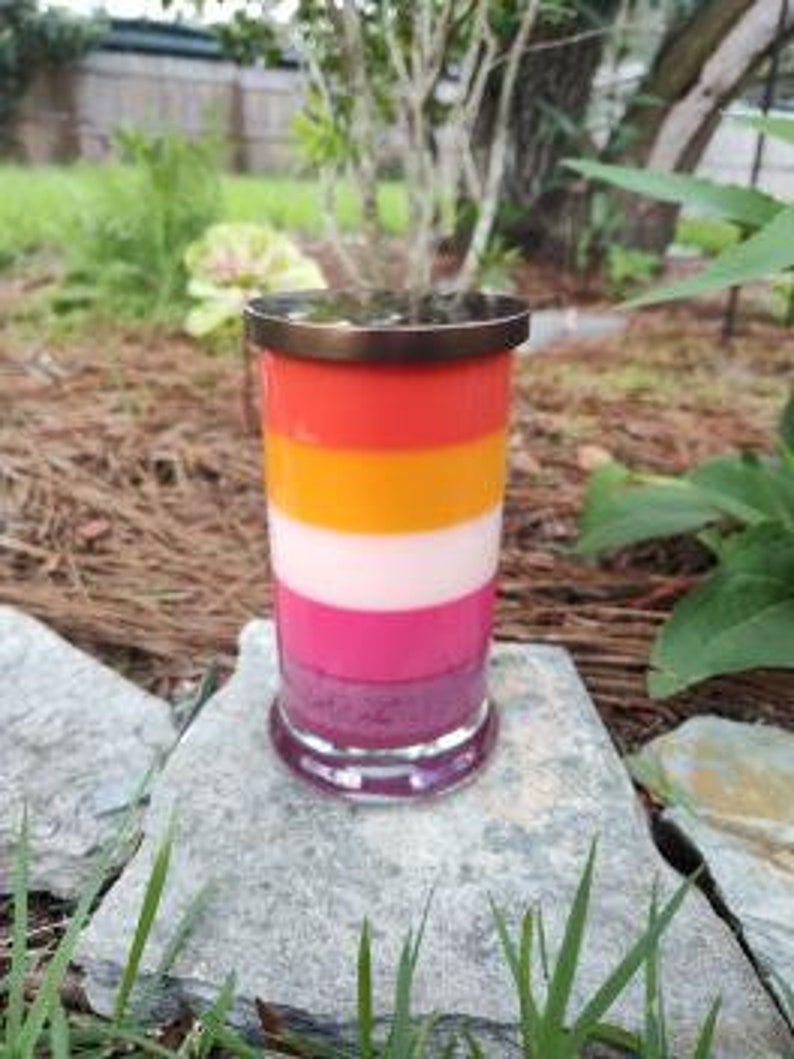 Op de afbeelding: Een gelaagde kaars in een glazen pot met een metalen deksel. De kaars is in de kleuren van de lesbische pride-vlag: roze, wit, oranje en rood.