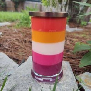 Op de afbeelding: Een gelaagde kaars in een glazen pot met een gouden deksel. De kaars is gelaagd in tinten rood, oranje, geel, wit, roze en paars.