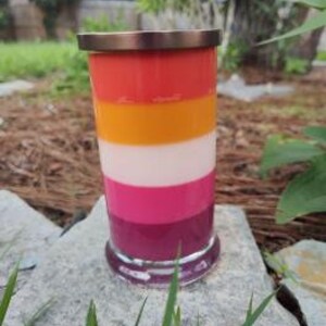 Op de afbeelding: Een gelaagde kaars in een glazen pot met een koperen deksel. De kaars is gelaagd in tinten rood, oranje, wit, roze en paars.