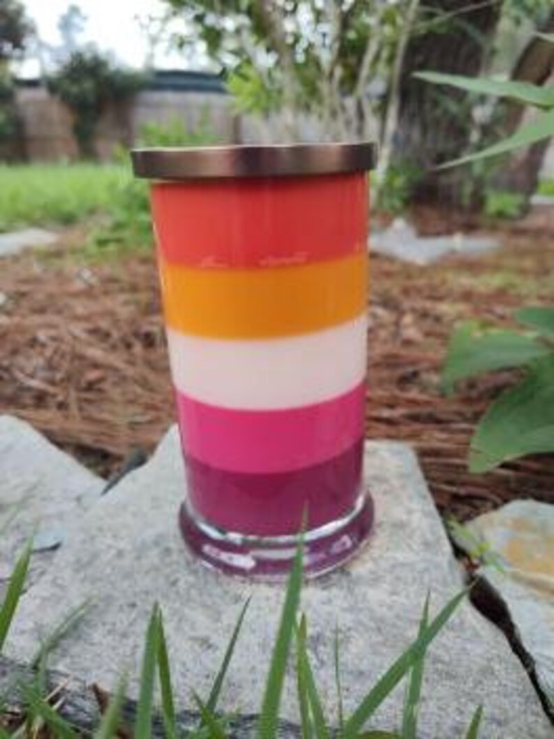 Op de afbeelding: Een gelaagde kaars in een glazen pot met een koperen deksel. De kaars is gelaagd in tinten rood, oranje, wit, roze en paars.