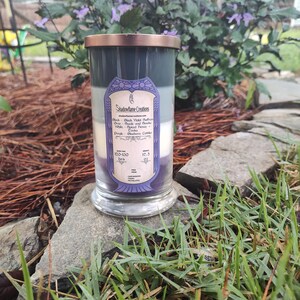 May include: A three-layered candle in a glass jar with a copper lid. The candle is labeled "Shadowflame Creations" and lists the scents of each layer: Black Violet Saffron, Spiced Honey, and Blueberry Cobbler. The candle has a burn time of 120-130 hours and weighs 17.5 oz.