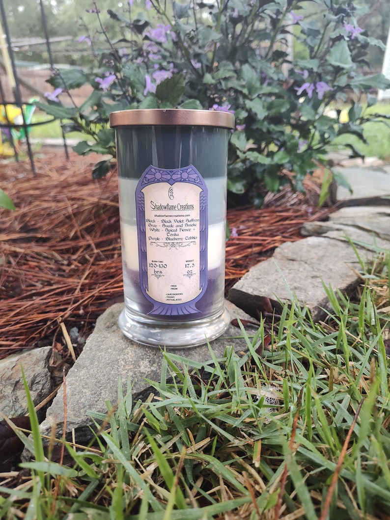 May include: A three-layered candle in a glass jar with a copper lid. The candle is labeled "Shadowflame Creations" and lists the scents of each layer: Black Violet Saffron, Spiced Honey, and Blueberry Cobbler. The candle has a burn time of 120-130 hours and weighs 17.5 oz.