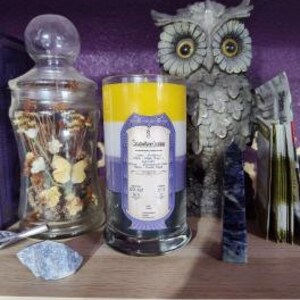 May include: A three-layered candle with yellow, white, and purple wax. The candle has a label that reads "Soothe Your Soul" and includes a list of essential oils. The candle is sitting on a wooden shelf with other decorative items.