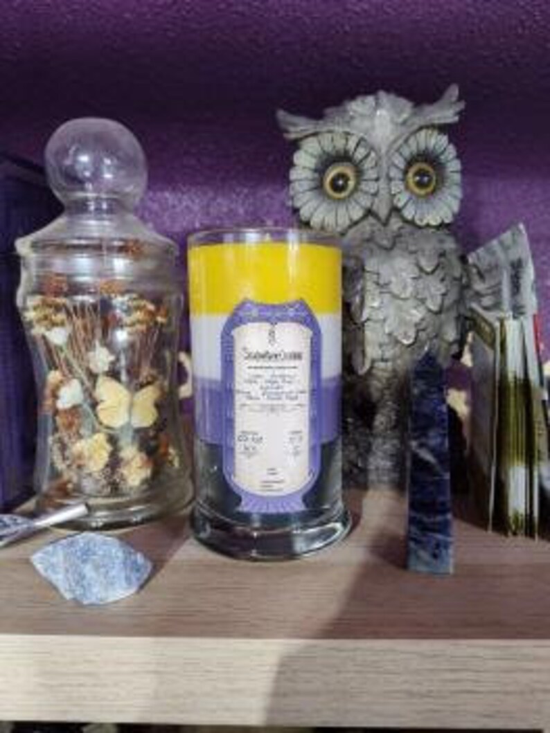 May include: A three-layered candle with yellow, white, and purple wax. The candle has a label that reads "Soothe Your Soul" and includes a list of essential oils. The candle is sitting on a wooden shelf with other decorative items.