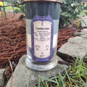 May include: A layered candle in a clear glass jar with a copper lid. The candle is divided into four sections: black, gray, white, and purple. The label on the jar reads "Shadowflame Creations" and lists the scents of each layer: Black Violet Saffron, Suede and Smoke, Spiced Honey + Tonka, and Blueberry Cobbler. The burn time is listed as 120-130 hours and the weight is 17.5 ounces. The candle is free from carcinogens, terns, and phthalates.