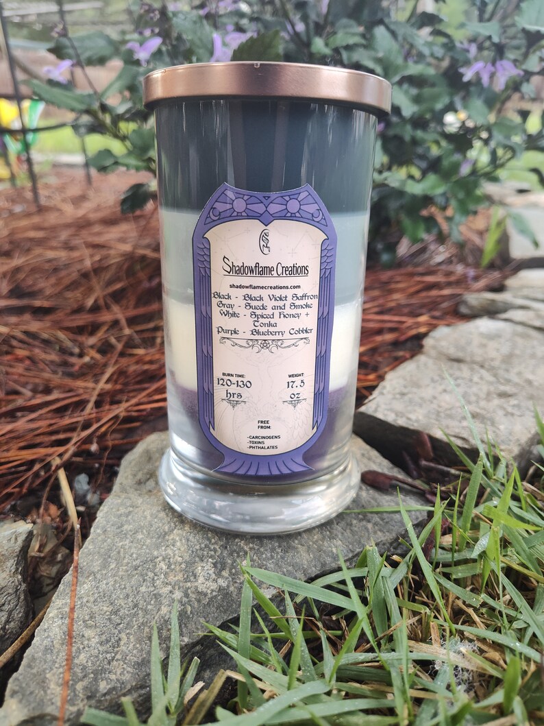 May include: A layered candle in a clear glass jar with a copper lid. The candle is divided into four sections: black, gray, white, and purple. The label on the jar reads "Shadowflame Creations" and lists the scents of each layer: Black Violet Saffron, Suede and Smoke, Spiced Honey + Tonka, and Blueberry Cobbler. The burn time is listed as 120-130 hours and the weight is 17.5 ounces. The candle is free from carcinogens, terns, and phthalates.