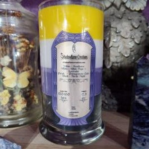 May include: A three-layered candle in a glass jar with a purple, white, and yellow color scheme. The label reads "Stoneflower Creations" and lists the scents as "Lavender, Cedarwood, Sage, Patchouli, Lavender, Black, Herbs, Herbs".