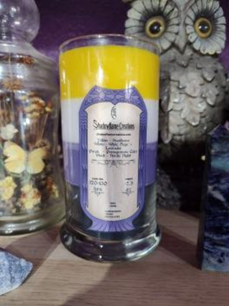 May include: A three-layered candle in a glass jar with a purple, white, and yellow color scheme. The label reads "Stoneflower Creations" and lists the scents as "Lavender, Cedarwood, Sage, Patchouli, Lavender, Black, Herbs, Herbs".