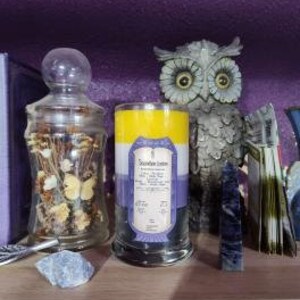 May include: A three-layered candle with a yellow, purple, and blue color scheme. The candle is in a clear glass jar with a black base. The label on the candle reads "Scentsation Station" and includes a list of ingredients.