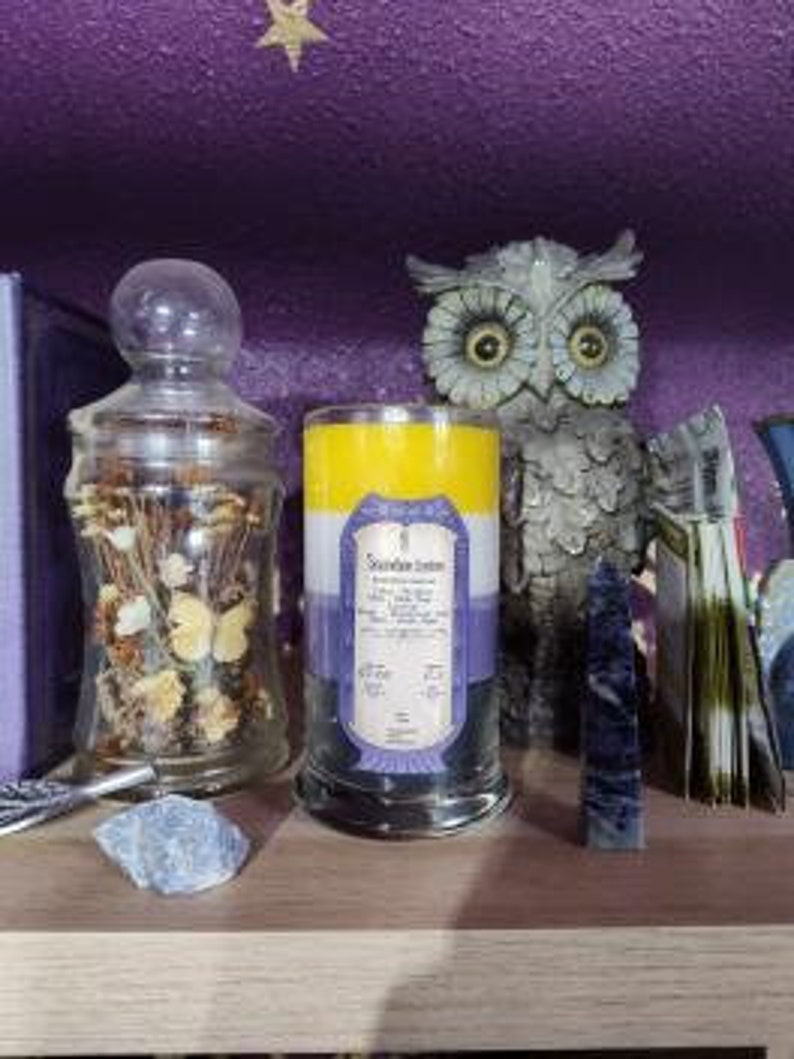 May include: A three-layered candle with a yellow, purple, and blue color scheme. The candle is in a clear glass jar with a black base. The label on the candle reads "Scentsation Station" and includes a list of ingredients.