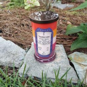 Op de afbeelding: Een meerkleurige kaars in een glazen pot met een metalen deksel. De kaars is gelaagd met rode, oranje, gele, witte en blauwe was. Het etiket op de kaars luidt "Chakra Crystal Candle" en somt de zeven chakra's op.