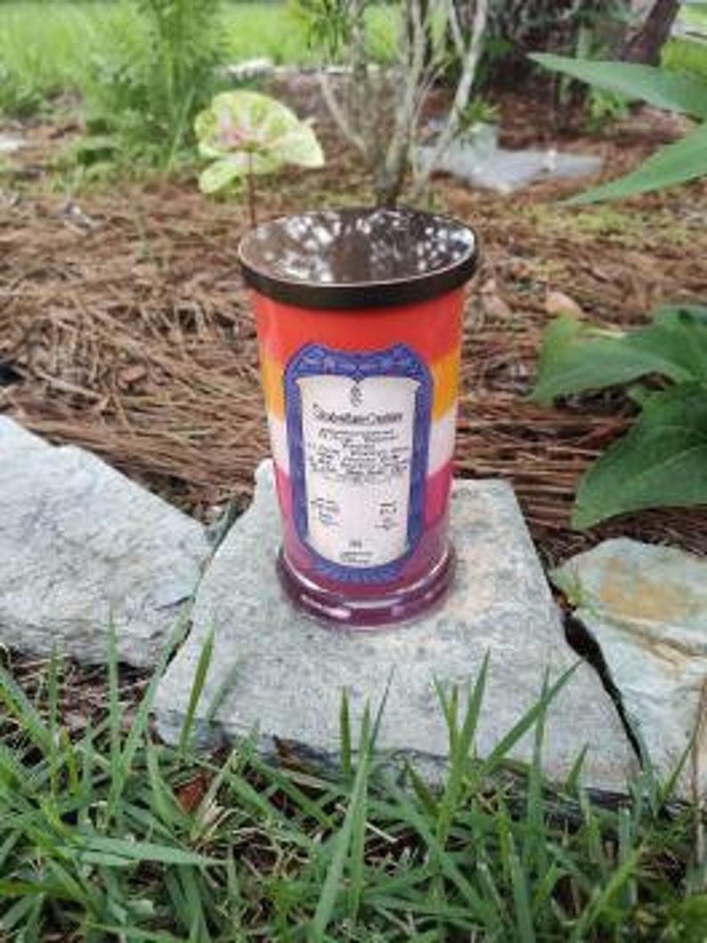 Op de afbeelding: Een meerkleurige kaars in een glazen pot met een metalen deksel. De kaars is gelaagd met rode, oranje, gele, witte en blauwe was. Het etiket op de kaars luidt "Chakra Crystal Candle" en somt de zeven chakra's op.