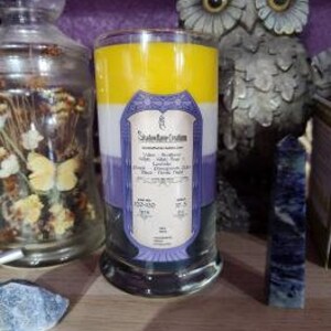 May include: A three-layered candle in a clear glass jar with a purple, white, and yellow color scheme. The candle has a label that reads "Sacred Space Candles" and lists the scents "Lavender, Butterfly Pea Flower, and White Tea".