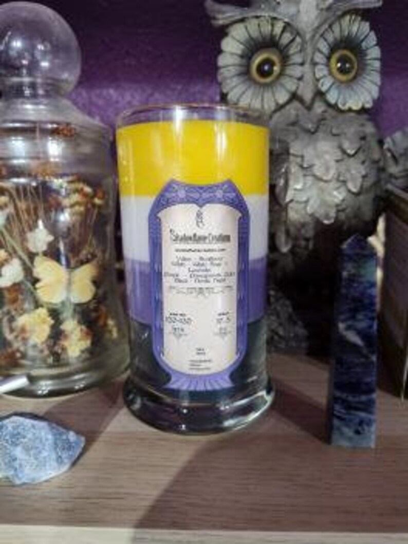 May include: A three-layered candle in a clear glass jar with a purple, white, and yellow color scheme. The candle has a label that reads "Sacred Space Candles" and lists the scents "Lavender, Butterfly Pea Flower, and White Tea".