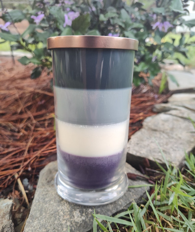 May include: A layered candle in a clear glass jar with a copper lid. The candle has four layers of wax: dark gray, light gray, white, and purple.
