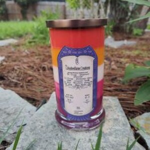 Op de afbeelding: Een gelaagde kaars in een glazen pot met een gouden deksel. De kaars is rood, oranje, geel en wit en heeft de tekst "Stonefire Creations" aan de zijkant. De kaars staat op een bed van groen gras.