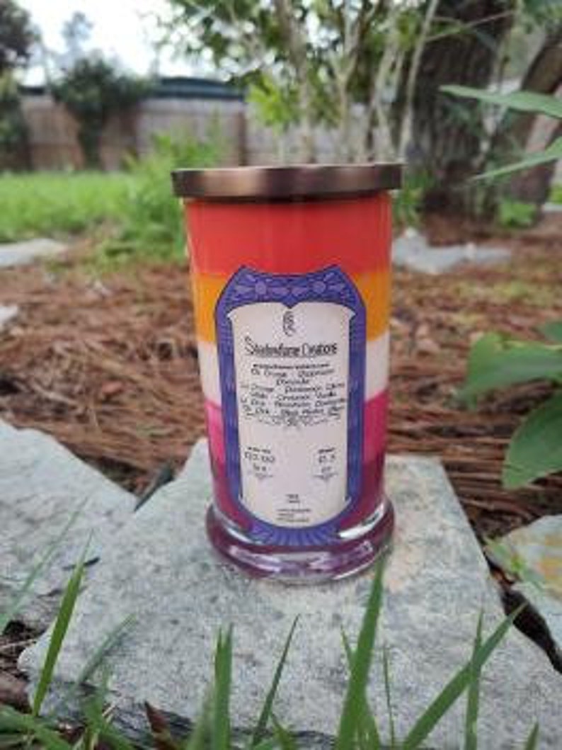 Op de afbeelding: Een gelaagde kaars in een glazen pot met een gouden deksel. De kaars is rood, oranje, geel en wit en heeft de tekst "Stonefire Creations" aan de zijkant. De kaars staat op een bed van groen gras.