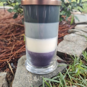 May include: A layered candle in a clear glass jar with a copper lid. The candle is made of four layers of wax: dark gray, light gray, white, and purple.