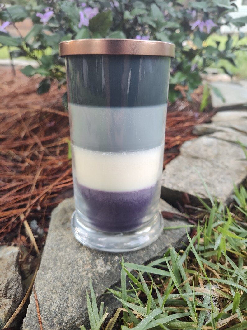 May include: A layered candle in a clear glass jar with a copper lid. The candle is made of four layers of wax: dark gray, light gray, white, and purple.
