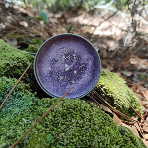 May include: A purple candle with a glittery finish, sitting on a bed of green moss. The candle has a speckled appearance, with small white flecks throughout.