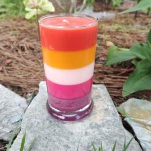 Op de afbeelding: Een gelaagde kaars in een glazen pot met een rood, oranje, wit en roze kleurenschema. De kaars staat op een grijze steen.