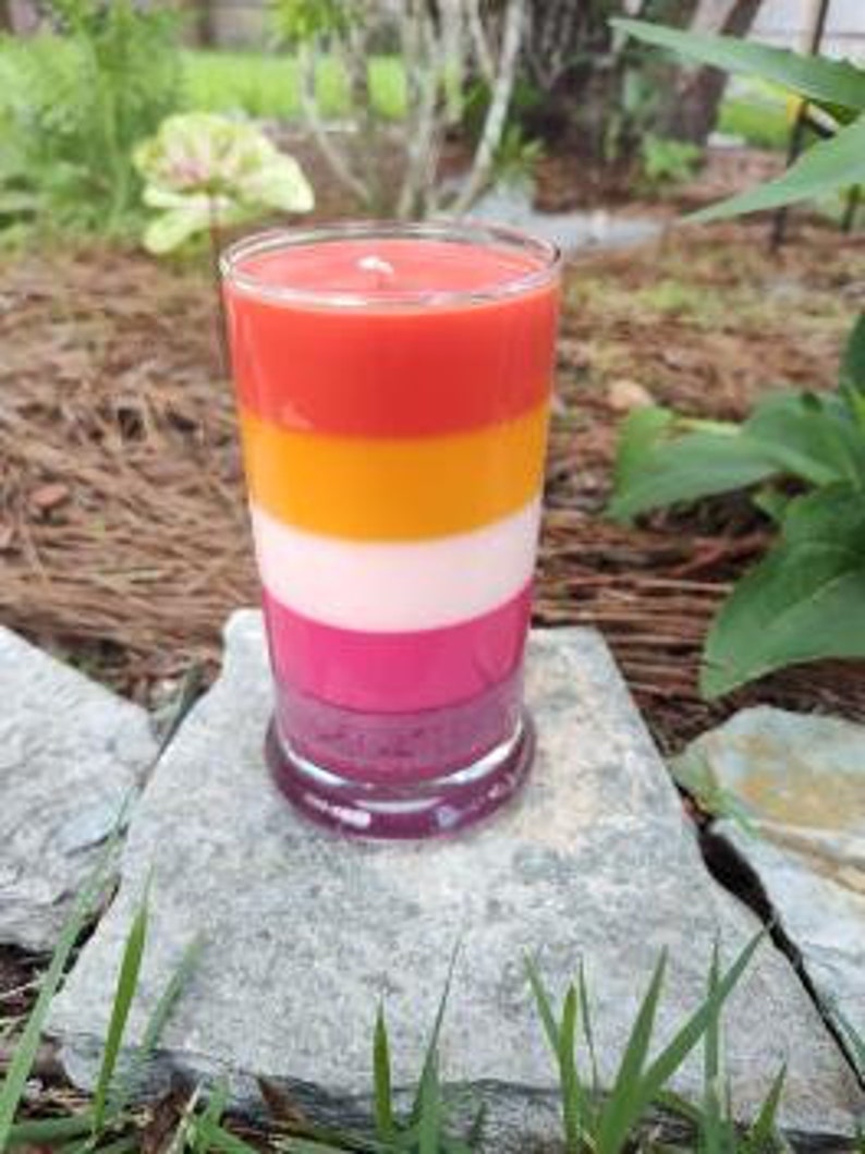 Op de afbeelding: Een gelaagde kaars in een glazen pot met een rood, oranje, wit en roze kleurenschema. De kaars staat op een grijze steen.