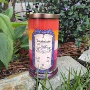 Op de afbeelding: Een meerkleurige kaars in een glazen pot met een koperen deksel. De kaars is gelaagd in tinten rood, oranje, geel en roze. Het etiket op de pot vermeldt "Smoldering Creations" en geeft de geur van de kaars aan als "The Queen's Garden".