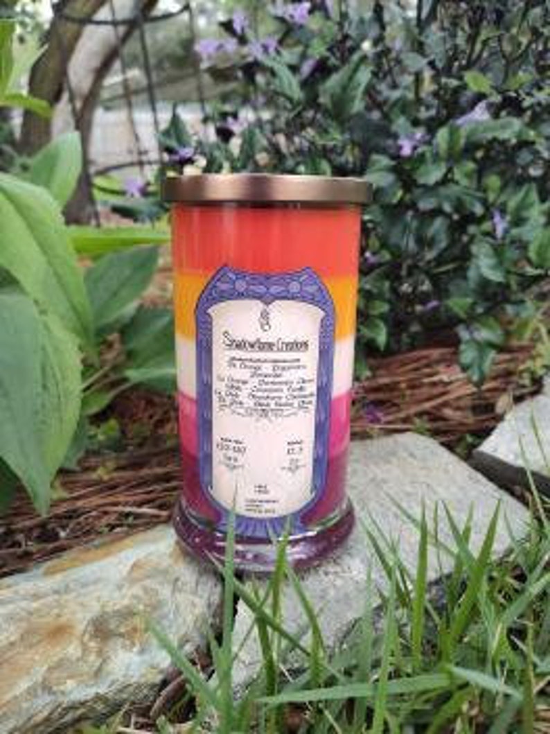 Op de afbeelding: Een meerkleurige kaars in een glazen pot met een koperen deksel. De kaars is gelaagd in tinten rood, oranje, geel en roze. Het etiket op de pot vermeldt "Smoldering Creations" en geeft de geur van de kaars aan als "The Queen's Garden".