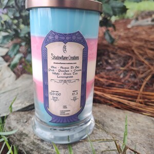 May include: A layered candle in a glass jar with a gold lid. The candle is blue, pink, and white, and has a label that says "Shadowflame Creations" and lists the scents: "Blue - Acqua Di Gio, Pink - Peaches + Cream, White - Green Tea, Lemongrass". The label also states the burn time is 120-130 hours and the weight is 17.5 oz.