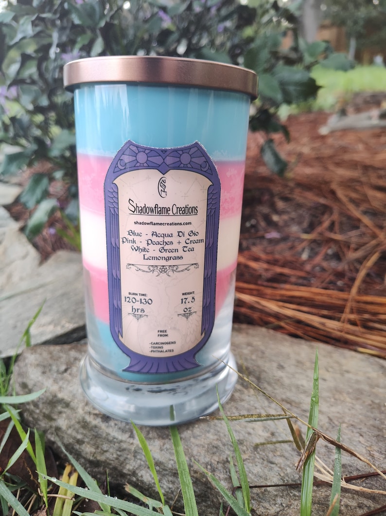May include: A layered candle in a glass jar with a gold lid. The candle is blue, pink, and white, and has a label that says "Shadowflame Creations" and lists the scents: "Blue - Acqua Di Gio, Pink - Peaches + Cream, White - Green Tea, Lemongrass". The label also states the burn time is 120-130 hours and the weight is 17.5 oz.