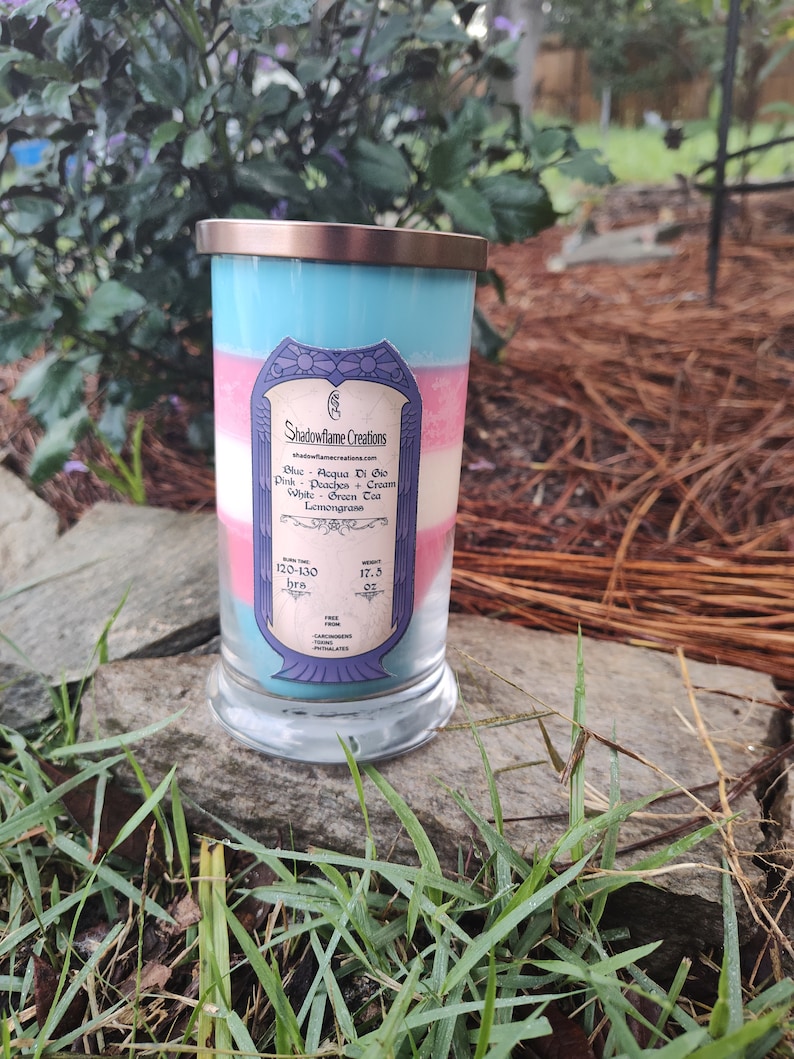May include: A glass candle jar with a copper lid. The candle is layered with blue, pink, and white wax. The label reads "Shadowflame Creations" and lists the scents: "Aqua Di Gio, Peaches & Cream, Green Tea, Lemongrass". The burn time is listed as 120-130 hours.