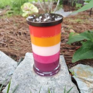 Op de afbeelding: Een gelaagde kaars in een bruine glazen pot met een kleurenschema van rood, oranje, wit, roze en paars.