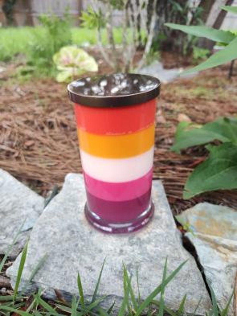 Op de afbeelding: Een gelaagde kaars in een bruine glazen pot met een kleurenschema van rood, oranje, wit, roze en paars.
