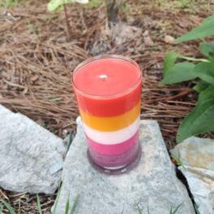 Op de afbeelding: Een gelaagde kaars in een glazen pot. De kaars is roze, wit, geel en rood. De kaars staat op een steen.
