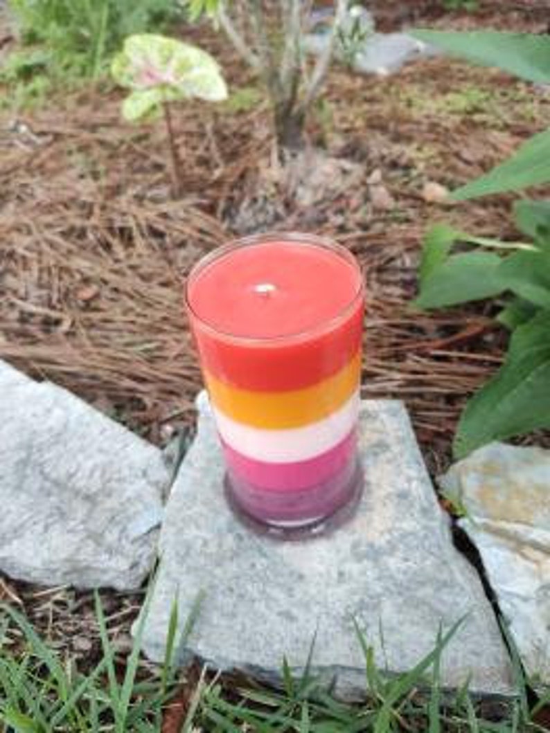Op de afbeelding: Een gelaagde kaars in een glazen pot. De kaars is roze, wit, geel en rood. De kaars staat op een steen.
