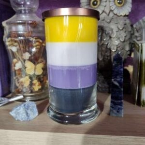 May include: A layered candle with yellow, white, purple, and gray wax in a clear glass jar with a gold lid. The candle is sitting on a wooden shelf.