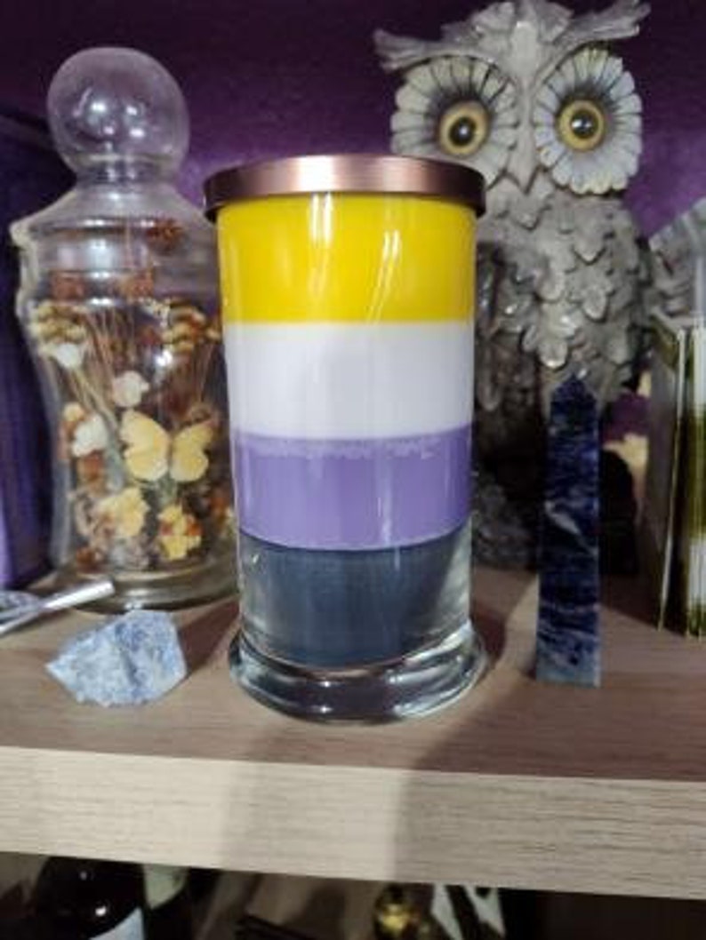 May include: A layered candle with yellow, white, purple, and gray wax in a clear glass jar with a gold lid. The candle is sitting on a wooden shelf.