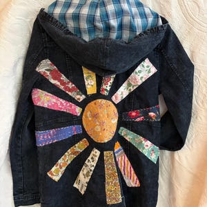 May include: A dark wash denim jacket with a hood lined in blue and white plaid fabric. The back features a sun-shaped appliqué made from a central yellow floral circle surrounded by radiating strips of various colorful floral and patterned fabrics.