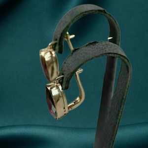 May include: A pair of gold-toned earrings featuring oval-shaped, deep red gemstones. The earrings are designed with a hinged lever-back closure mechanism, showcasing a polished metal frame that secures the faceted stones in a vertical orientation.