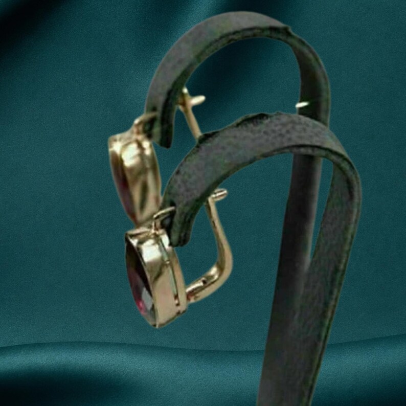 May include: A pair of gold-toned earrings featuring oval-shaped, deep red gemstones. The earrings are designed with a hinged lever-back closure mechanism, showcasing a polished metal frame that secures the faceted stones in a vertical orientation.