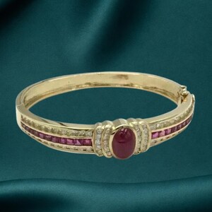 May include: A gold hinged bangle bracelet featuring a central oval cabochon ruby flanked by two rows of small round diamonds. The band is accented with a channel-set row of square-cut rubies and a row of small round diamonds on each side.