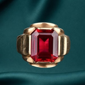 May include: A gold ring featuring a large, rectangular, emerald-cut red gemstone set in the center. The band has a wide, smooth, polished gold finish with tiered, architectural side details that frame the vibrant red stone securely.