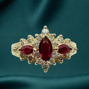 May include: A gold ring featuring a central marquise-cut red gemstone flanked by two smaller marquise-cut red gemstones, all surrounded by a cluster of round white diamonds set in a symmetrical arrangement on a polished gold band.