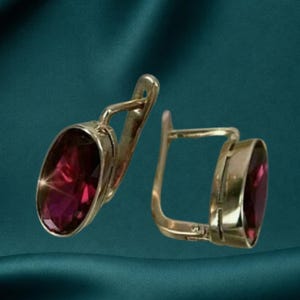 May include: A pair of gold-toned earrings featuring oval-shaped, deep magenta-coloured faceted stones. Each earring is designed with a hinged lever-back closure, securely holding the polished metal setting that frames the vibrant, translucent gemstone.