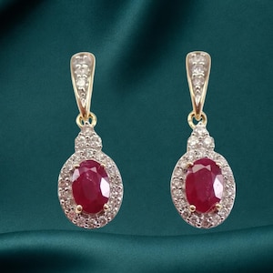 May include: A pair of drop earrings featuring oval-cut red rubies set in gold. Each ruby is surrounded by a halo of small white diamonds, with additional diamonds set along the gold bale at the top of each earring, creating a symmetrical design.